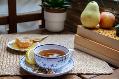 Balkondaki ahşap masa, okunacak kitaplar, bir fincan doğal çay, çaydanlık, çiftlikten gelen organik bal, taze yeşil çay yaprakları ve organik meyveler, arka planda binalar arasındaki küçük park manzarası, dinlenmek için küçük vaha.