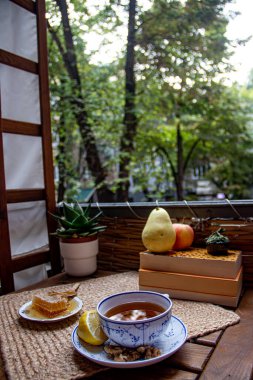 Balkondaki ahşap masa, okunacak kitaplar, bir fincan doğal çay, çaydanlık, çiftlikten gelen organik bal, taze yeşil çay yaprakları ve organik meyveler, arka planda binalar arasındaki küçük park manzarası, dinlenmek için küçük vaha.