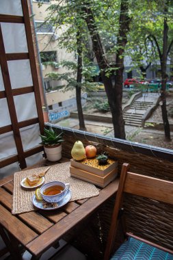Balkondaki ahşap masa, okunacak kitaplar, bir fincan doğal çay, çaydanlık, çiftlikten gelen organik bal, taze yeşil çay yaprakları ve organik meyveler, arka planda binalar arasındaki küçük park manzarası, dinlenmek için küçük vaha.