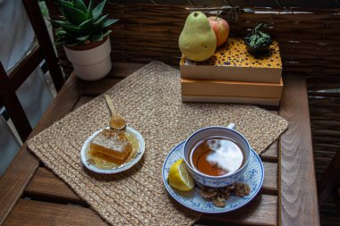 Balkondaki ahşap masa, okunacak kitaplar, bir fincan doğal çay, çaydanlık, çiftlikten gelen organik bal, taze yeşil çay yaprakları ve organik meyveler, arka planda binalar arasındaki küçük park manzarası, dinlenmek için küçük vaha.