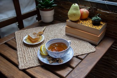 Balkondaki ahşap masa, okunacak kitaplar, bir fincan doğal çay, çaydanlık, çiftlikten gelen organik bal, taze yeşil çay yaprakları ve organik meyveler, arka planda binalar arasındaki küçük park manzarası, dinlenmek için küçük vaha.