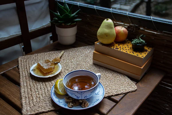 Morning setup on wooden table at balcony, books to read, cup of natural tea, teapot, organic honey from farm, fresh green tea leaves and organic fruits, in background nice view on small park between buildings in city, small oasis for relaxation