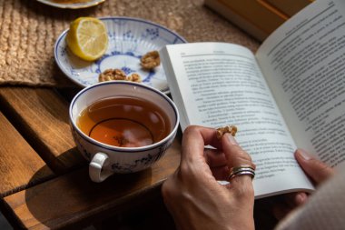 Balkondaki ahşap masa, okunacak kitaplar, bir fincan doğal çay, çaydanlık, çiftlikten gelen organik bal, taze yeşil çay yaprakları ve organik meyveler, arka planda binalar arasındaki küçük park manzarası, dinlenmek için küçük vaha.