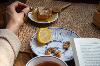 Balkondaki ahşap masa, okunacak kitaplar, bir fincan doğal çay, çaydanlık, çiftlikten gelen organik bal, taze yeşil çay yaprakları ve organik meyveler, arka planda binalar arasındaki küçük park manzarası, dinlenmek için küçük vaha.