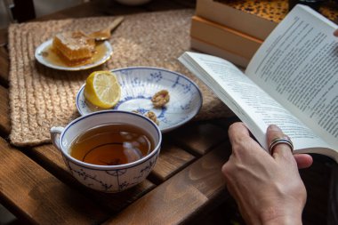 Balkondaki ahşap masa, okunacak kitaplar, bir fincan doğal çay, çaydanlık, çiftlikten gelen organik bal, taze yeşil çay yaprakları ve organik meyveler, arka planda binalar arasındaki küçük park manzarası, dinlenmek için küçük vaha.