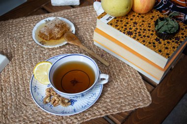 Balkondaki ahşap masa, okunacak kitaplar, bir fincan doğal çay, çaydanlık, çiftlikten gelen organik bal, taze yeşil çay yaprakları ve organik meyveler, arka planda binalar arasındaki küçük park manzarası, dinlenmek için küçük vaha.