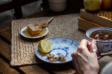 Balkondaki ahşap masa, okunacak kitaplar, bir fincan doğal çay, çaydanlık, çiftlikten gelen organik bal, taze yeşil çay yaprakları ve organik meyveler, arka planda binalar arasındaki küçük park manzarası, dinlenmek için küçük vaha.