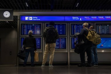 Gece geç saatlerde havaalanındaki bir bilgi panosunun önünde durup aktarma bölgesini kontrol ettikten sonra bağlantı bilgilerini arayan ve düzlükte uçmayı bekleyen yolcular.