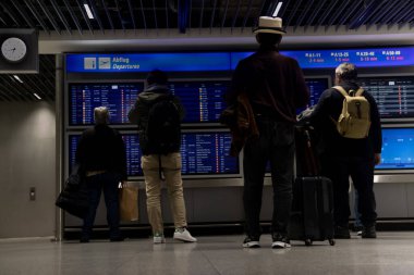 Gece geç saatlerde havaalanındaki bir bilgi panosunun önünde durup aktarma bölgesini kontrol ettikten sonra bağlantı bilgilerini arayan ve düzlükte uçmayı bekleyen yolcular.