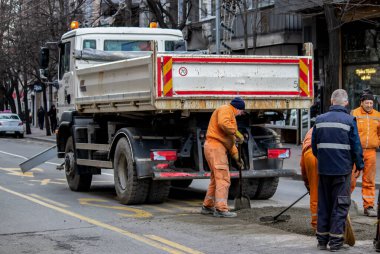 Turuncu üniformalı ve koruma ekipmanlı işçiler büyük şehir merkezindeki hasarlı yolu onarıyor ve onarıyor. Ağır makineler ve kirli işler.