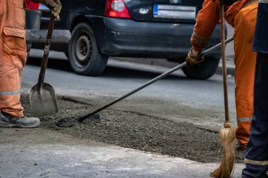 Turuncu üniformalı ve koruma ekipmanlı işçiler büyük şehir merkezindeki hasarlı yolu onarıyor ve onarıyor. Ağır makineler ve kirli işler.