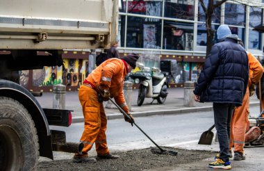 Turuncu üniformalı ve koruma ekipmanlı işçiler büyük şehir merkezindeki hasarlı yolu onarıyor ve onarıyor. Ağır makineler ve kirli işler.