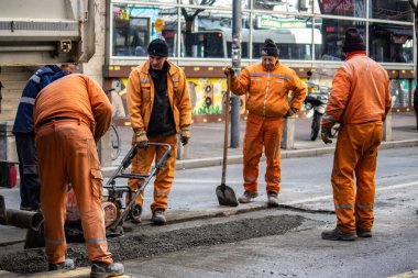 Turuncu üniformalı ve koruma ekipmanlı işçiler büyük şehir merkezindeki hasarlı yolu onarıyor ve onarıyor. Ağır makineler ve kirli işler.