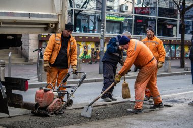 Turuncu üniformalı ve koruma ekipmanlı işçiler büyük şehir merkezindeki hasarlı yolu onarıyor ve onarıyor. Ağır makineler ve kirli işler.