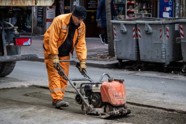 Turuncu üniformalı ve koruma ekipmanlı işçiler büyük şehir merkezindeki hasarlı yolu onarıyor ve onarıyor. Ağır makineler ve kirli işler.