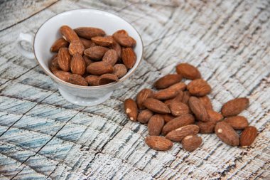 Ahşap masada kavrulmuş organik kahverengi badem tohumu ve beyaz porselen bardakta biraz badem yakın.