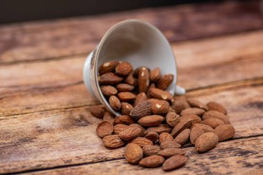 Ahşap masada kavrulmuş organik kahverengi badem tohumu ve beyaz porselen bardakta biraz badem yakın.