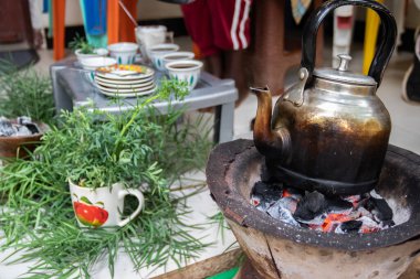 A jebena sits on a grass-covered table with small, handle-less cups arranged around it. Freshly roasted coffee beans, a mortar and pestle, and incense smoke create a traditional Ethiopian coffee ceremony setup.