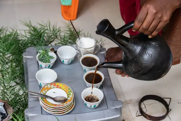 stock image A jebena sits on a grass-covered table with small, handle-less cups arranged around it. Freshly roasted coffee beans, a mortar and pestle, and incense smoke create a traditional Ethiopian coffee ceremony setup.