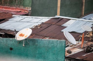 Eski, paslı uydu çanakları yerleşim yerinin çatılarını süslüyor, eski teknolojiyi yansıtıyor ve toplumdaki yıpranmış altyapıyı yansıtıyor.
