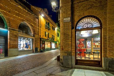 ALBA, İtalya - 24 Kasım 2020: Kafenin aydınlatılmış penceresi, eski Alba kasabasındaki tarihi binalar boyunca yılbaşı aydınlatmasıyla dar kaldırım taşı caddesi.