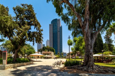 Tel Aviv 'deki Sarona pazarının arka planında mavi gökdelenler ve şehir parkı.