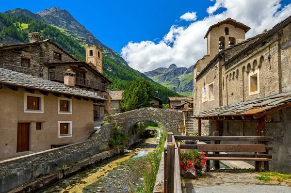 アルパインクリークは中世の石造りの橋の下を流れ 北イタリアのピエモンテ州のチアナーレの小さな村を背景に山として古い家や教区教会の間を流れている — ストック写真