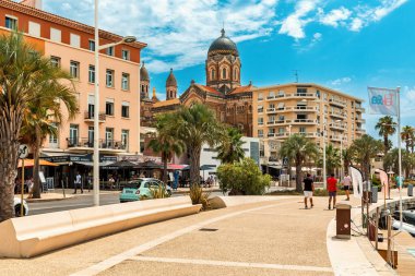 Saint-Rafael, FRANCE - 29 Temmuz 2022: Saint-Raphael şehrinin arka planında, mavi gökyüzü altında gezinti güvertesi, binalar ve katedral - ünlü tatil beldesi ve popüler seyahat merkezi.