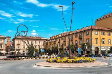 ALBA, ITALY - 29 Nisan 2023: Lamppost, Alba 'nın arka planındaki tarihi binalar arasında modern heykel ile merkezi kare olarak çiçeklerle süslenmiş döner kavşak üzerinde, ünlü seyahat merkezi Piedmont kasabası.