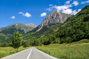 Yol yeşil tarlalardan ve yamaçlardan geçer. Mavi gökyüzünün altındaki dağ sırtı gibi. İtalya, Piedmont 'ta arka planda..