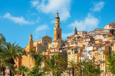 Saint Michel Archange Bazilikası, açık mavi gökyüzü altındaki renkli binalarla çevrili uzun sarı çan kulesiyle eski Fransa 'nın Menton kasabasında güneşli bir günde.