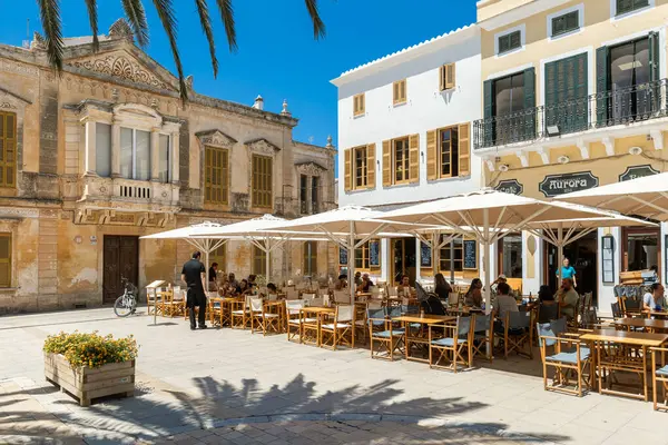 CIUTADELLA, SPAIN - 11 Temmuz 2024: İnsanlar Ciutadella 'da tarihi mimari ile çevrili küçük bir kasaba meydanındaki büyük beyaz şemsiyelerin altında masalarda oturuyorlar - Menorca' da eski bir şehir, popüler seyahat merkezi.