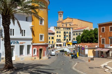 MAHON, SPAIN - 10 HAZİRAN 2024: Renkli binalar, palmiye ağaçları ve Mahon 'da mavi gökyüzünün altında bir kilise kulesi olan bir sokak manzarası - Balear Adaları' ndaki tarihi Akdeniz kasabası, ünlü seyahat merkezi.