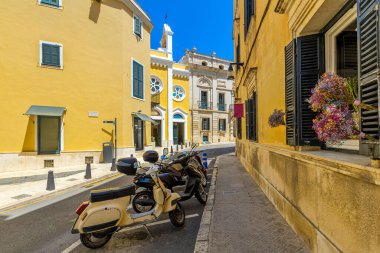 İspanya 'nın Balear Adaları' ndaki eski Mahon kasabasında, canlı sarı binalar ve tarihi mimari zemine parketmiş scooterlarla dar bir sokak..