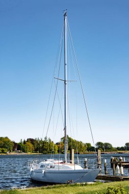 Kürklü yelkenleri olan beyaz yelkenli güneşli bir günde çimenli sahilde ahşap rıhtımla demirli..