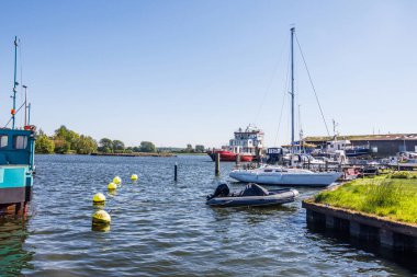 Güneşli bir günde sarı şamandıralı bir limana demirlemiş beyaz yelkenli ve siyah motorlu tekne..