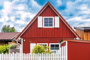 Beyaz pencere panjurları, beyaz bahçe çitleri ve bulutlu mavi gökyüzü olan büyüleyici kırmızı ahşap bir ev.