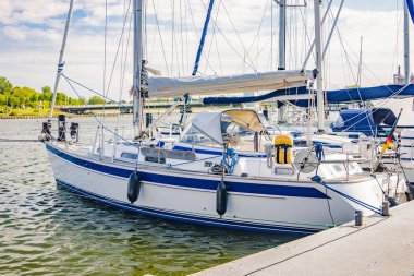 Güneşli bir günde limandaki bir rıhtıma demirlemiş beyaz yelkenli yat, deniz yaşamı ve boş zaman aktivitelerini gösteriyor.