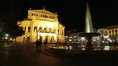 Aydınlanmış Alte Oper Frankfurt ve çeşmesinin yanında yürüyen turistler