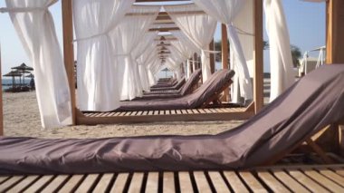 Güneş sandalyesi ve kumsalda beyaz perdeleri olan lüks bir salon. Tatil, rahatlama ve tatil konsepti. Yüksek kaliteli FullHD görüntüler