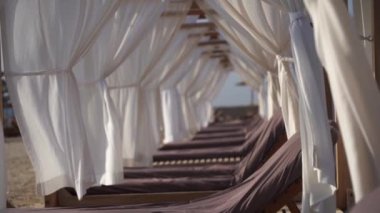 Güneş sandalyesi ve kumsalda beyaz perdeleri olan lüks bir salon. Tatil, rahatlama ve tatil konsepti. Yüksek kaliteli FullHD görüntüler
