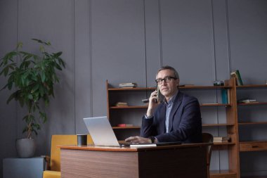 Senior businessman using his mobile phone and talking wih somebody while working laptop in the office.