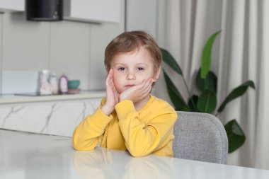 Pensive child boy with brown hair in yellow sweater sitting by the table on studio kitchen background