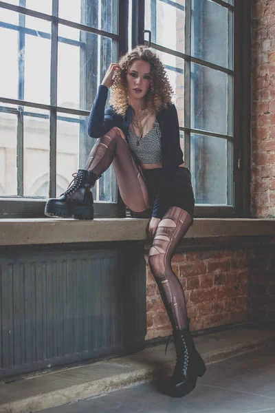 Beautiful glamorous grunge woman with torn tights and cutly hair sitting by the window on the windowsill