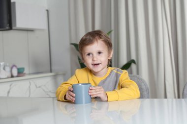 Sarı süveterli mutlu çocuk masanın yanında oturuyor ve elinde gri bardakla beyaz perde arkasında