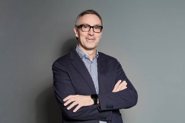 Gri arka planda duran takım elbiseli ve gözlüklü mutlu iş adamı gülümsüyor.