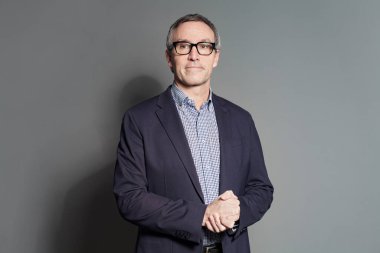 Olgun gri saçlı takım elbiseli ve gözlüklü iş adamı gri stüdyo arka planında duruyor.