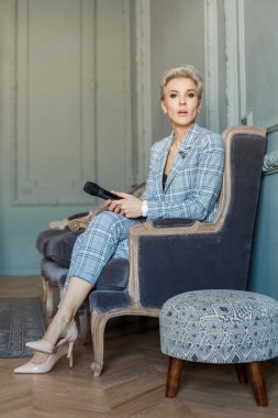 Mavi takım elbiseli, beyaz tepeli güzel mutlu kadın gazeteci kanepede oturuyor ve mikrofon tutuyor.