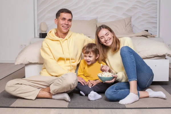 stock image Cute laughing smiling parents with child son sitting by the bed and eating popcorn