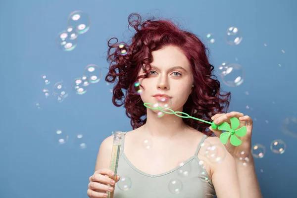 Woman with red curly hair blowing soap bubbles against blue studio wall background